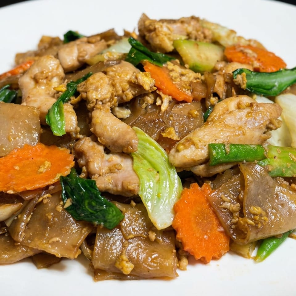 Stir-fried wide rice noodles with chicken, carrots, leafy greens, onions, and broccoli on a white plate.